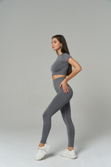 Athletic woman in gray sportswear stands sideways, showing fit physique. Long hair, poised posture, and white sneakers. Concept of fitness, healthy lifestyle, and fashion in sports