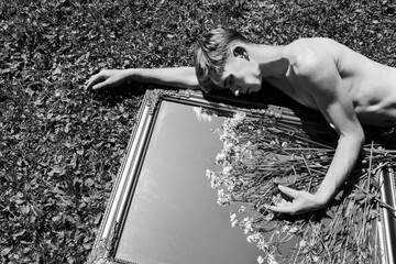 A shirtless young man lies on grass, embracing a large ornate mirror with flowers on it
