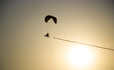 Paramotor silhouette flying across golden sky at sunset. Soft orange gradient background, minimalistic composition with warm light and peaceful evening atmosphere.