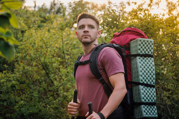 Attractive hiker with backpack and trekking poles in forest at sunset. Portrait of man hiker