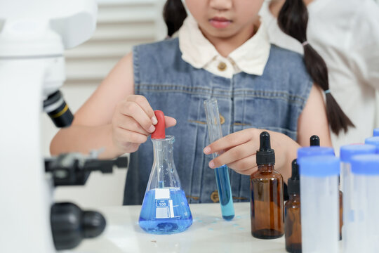 Closeup view of asian schoolgirl using pipette to drop blue liquid into test tube during science experiment in classroom with lab bottles and microscope on white science table