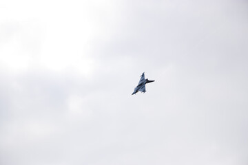 Military fighter jet in flight against a clear pale blue sky. Dark silhouette of aircraft with sharp angular wings in dynamic motion, minimalistic aerial view.

