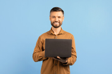 Confident young man smiling and holding laptop in hands. Handsome male using computer for remote...
