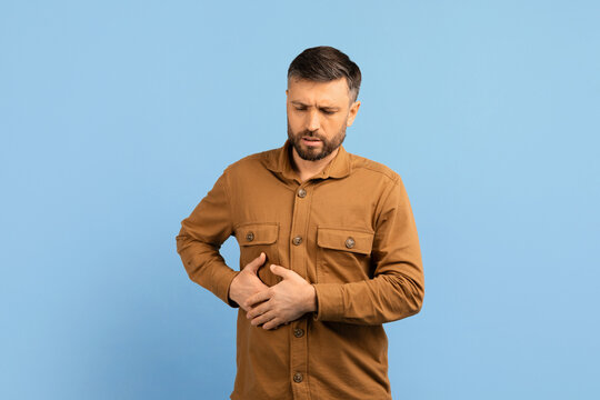 Man touching right side of his abdomen with painful expression. Studio concept for liver pain, gallbladder issue, or internal organ discomfort, sick man standing on blue background