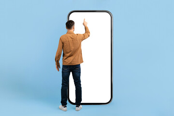 Rear view of young man interacting with large smartphone mockup. Blank screen design concept ideal...