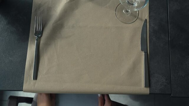 Top view of a grey table with a napkin, a glass and cutlery, where a woman sits and a waiter serves pasta on a white plate. Service and maintenance in the restaurant according to rules of etiquette