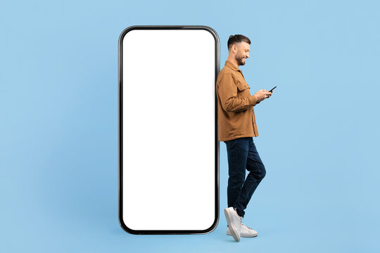 Smiling young man leaning on oversized blank smartphone with white screen and using mobile. Studio shot on blue background, perfect for technology, app promotion, or digital lifestyle themes - Powered by Adobe