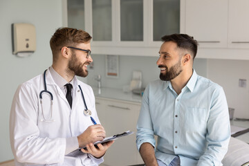 Positive male Caucasian physician talking to young Latin patient man on consultation meeting,...