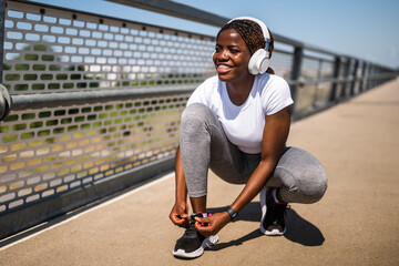 Motivated young sporty African-American woman tying her shoelaces on a city bridge before jogging,...