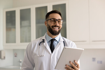 Cheerful young Latin doctor man in glasses holding tablet, standing in hospital office, looking...