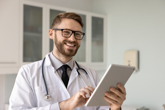 Happy young Caucasian doctor man in stylish eyeglasses and white coat typing on digital gadget, using tablet fir online medical communication, chatting with patients, looking away, smiling, laughing