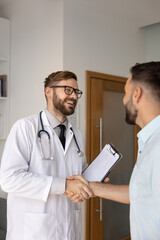 Positive young Caucasian doctor man wearing lab coat and glasses, shaking hands with patient,...