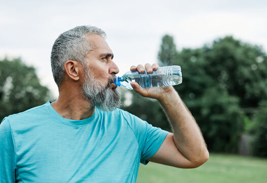 Smiling active mature mid aged coman jogging exercising and having fun and l taking a break drinking water in the park - Powered by Adobe