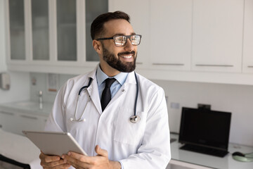 Happy handsome Hispanic doc in trendy glasses and white coat standing in clinic office with tablet,...