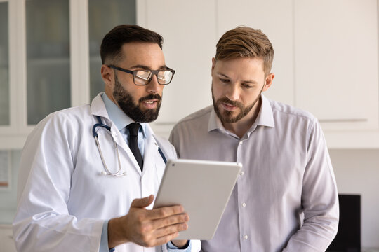 Serious Hispanic medical practitioner man and young patient looking at electronic medical report, examination results on tablet. Doctor explaining diagnosis, online prescription to man - Powered by Adobe