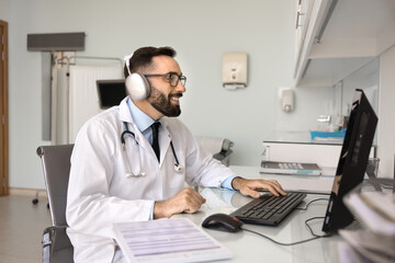 Happy friendly Hispanic doctor man in headphones and white lab coat talking to patient on video...