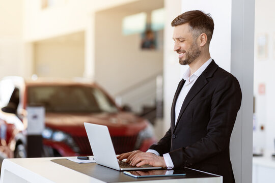 Professional car salesman working at laptop in dealership showroom. Handsome man preparing documents or checking inventory with a smile in bright, elegant workspace, copy space - Powered by Adobe