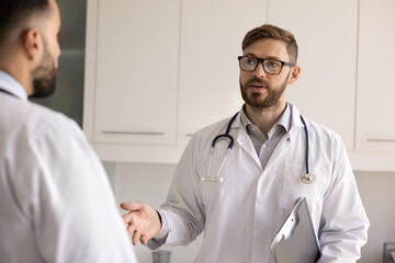 Serious young Caucasian doctor man in glasses and lab coat talking to male coworker in hospital...