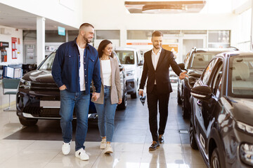 Car dealer explaining features of a vehicle to young couple at modern showroom. Smiling man and woman listening with interest and excitement during car purchase, full length