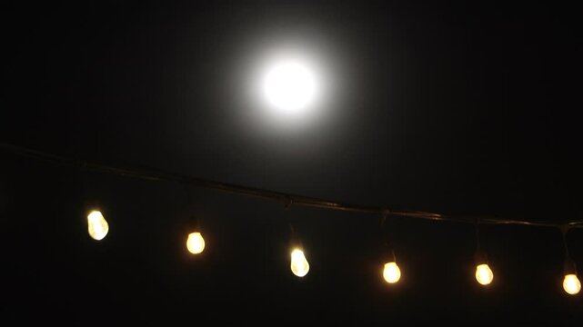 A chain of glowing light bulbs moving in the wind with a background of a shining full moon