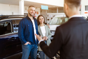 Happy young couple smiling while listening to car dealer in a showroom. Positive emotions and excitement during car buying process, selective focus to family