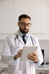 Serious busy Hispanic physician man in lab coat and stylish glasses typing on tablet, using digital...