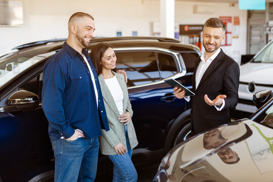 Car dealer showing car options to a young couple at a dealership center. Friendly consultation in auto showroom with smiling faces and modern vehicles in the background