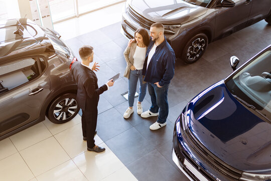 Top view of smiling couple talking to car salesman in a dealership. Family choosing auto in modern, open space showroom with multiple cars and confident communication