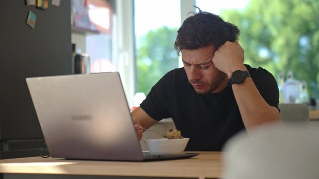Man sitting at kitchen table, eats oatmeal porridge with displeased look and pushed her plate away. Unhappy man eating tasteless breakfast in kitchen table with laptop