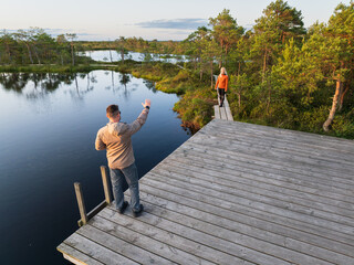 A couple of tourists on a walk in a picturesque bog: the man on a wooden pier waves in greeting to the woman on the trail.