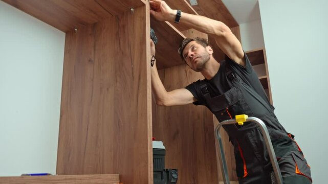 Man wearing work overalls assembling wooden wardrobe with drill and tools. Carpenter standing on step ladder, furniture installation process