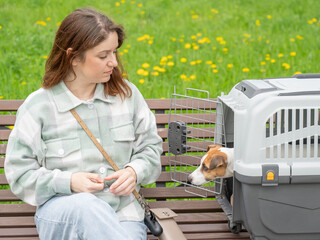 Caucasian woman traveling with dog. Owner sits on bench with Jack Russell Terrier in cage for safe...