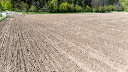 Aerial view of freshly plowed agricultural field with tire tracks, surrounded by lush green trees in spring, highlighting farmland's preparation for planting crops.