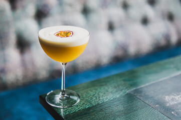 Close-up of a yellow cocktail with passion fruit garnish in a coupe glass, set on a green wooden...