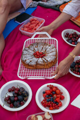 Summer picnic with fresh fruits and homemade cake. Hands serving dessert on a pink blanket outdoors. Cozy gathering in nature.