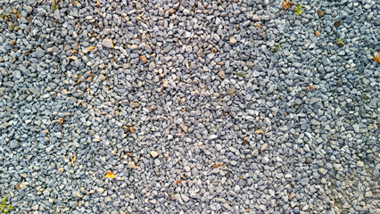 Close-up view of a gravel surface, showcasing various sizes of gray stones intermixed with small bits of dirt and green foliage. Suitable for landscaping or construction contexts.