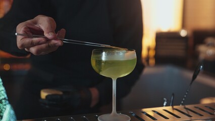 Cropped bartender garnishing caramelized citrus slice on cube ice in green Midori Sour cocktail served in stylish stemmed glass. Perfect for sophisticated bar settings christmas tree theme. Vinosity.