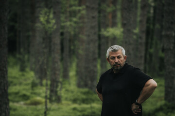A mature man with gray hair and beard stands with hands on hips in a dense forest, wearing a black shirt and gazing slightly to the side with a serious expression.