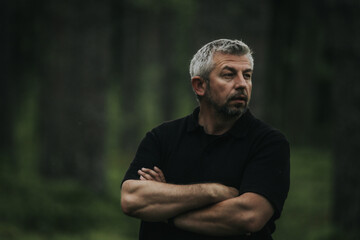 A man with gray hair and a beard stands with his arms crossed, looking up into a shady forest. He is wearing a black shirt, and his mood is contemplative and calm.