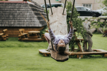 A happy girl in a lavender sweatshirt swings upside down on a rustic wooden swing in a grassy yard with wooden decor and parked cars in the background.