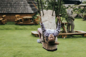 A happy girl in a lavender sweatshirt swings upside down on a rustic wooden swing in a grassy yard with wooden decor and parked cars in the background.