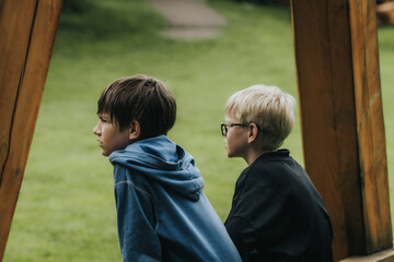 Two boys sit side by side under a wooden structure, thoughtfully observing a green lawn. An...