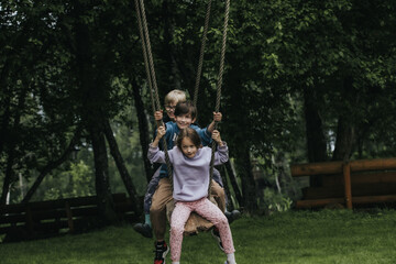 Three children sit close together on a wooden rope swing in a wooded area, laughing and playing...