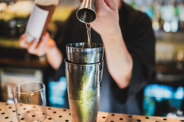 Bartender pouring liquid from jigger into cocktail shaker at bar counter, close-up of hands and bar tools during drink preparation in a modern nightlife setting