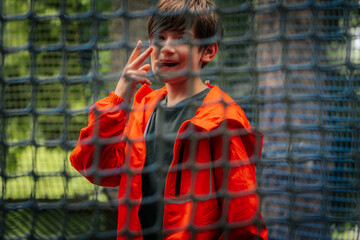 A cheerful boy in an orange jacket poses with a peace sign behind a blue safety net outdoors, in a playful atmosphere, surrounded by greenery.