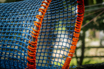 An empty blue-orange rope tunnel suspended in a wooded outdoor playground, forming a curved, mesh structure.