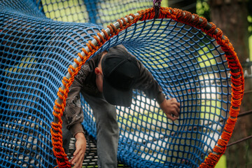 A little boy in a hat and hoodie climbs through a bright blue and orange rope tunnel at an outdoor playground.