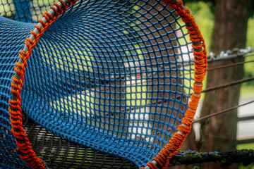 An empty blue-orange rope tunnel suspended in a wooded outdoor playground, forming a curved, mesh structure.