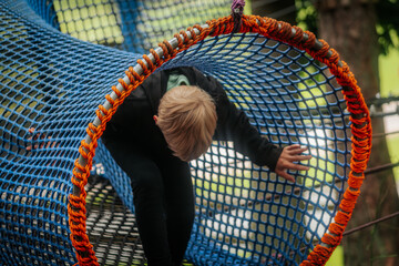 A small child in a black hoodie moves through a colorful rope tunnel made of blue and orange netting in a forest adventure park.