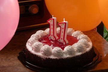 A decorated birthday cake with chocolate chips, whipped cream swirls, and number 11 candles sits on a wooden table, surrounded by colorful balloons and an antique radio.
